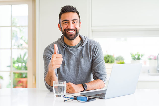 Handsome hispanic man working using computer laptop doing happy thumbs up gesture with hand. Approving expression looking at the camera with showing success.